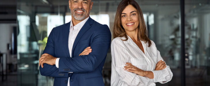 Portrait of smiling mature Latin or Indian business man and European business woman standing arms crossed in office. Two diverse colleagues, group team of confident professional business people.